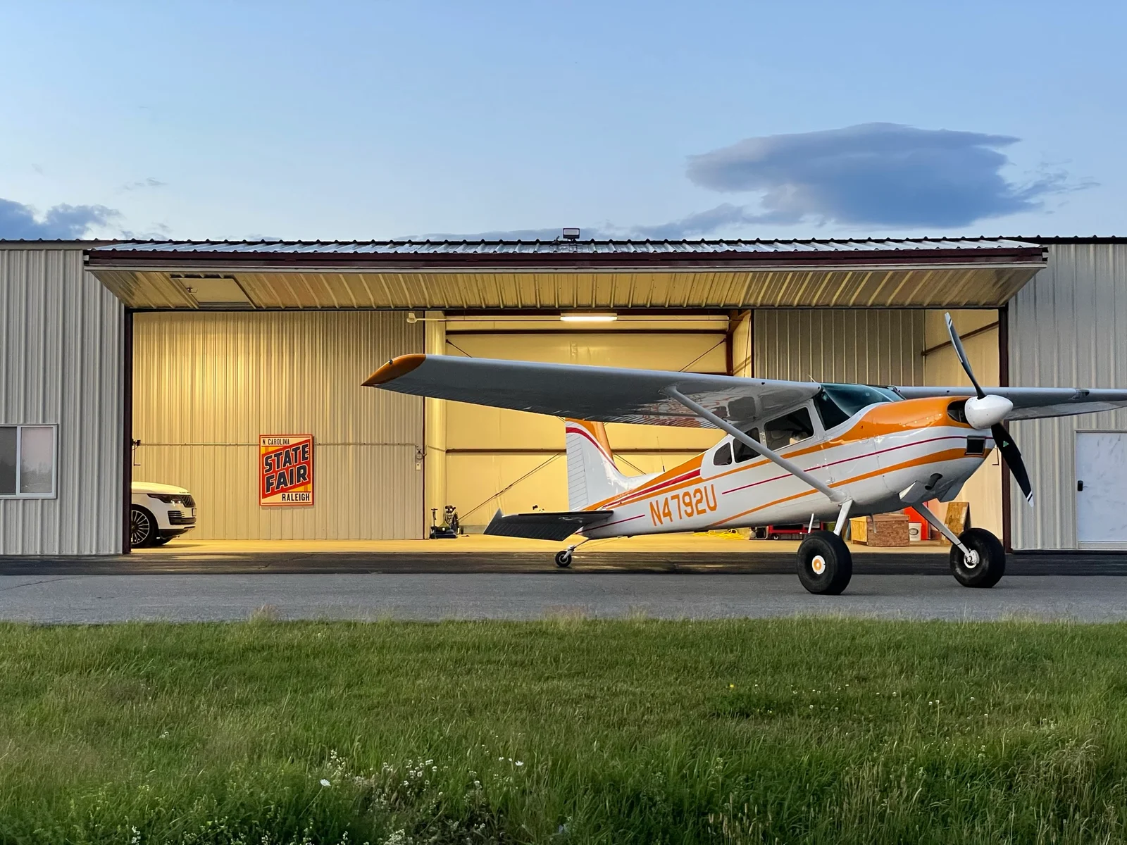Atlas’s Cessna 180 parked outside an open, warmly lit hangar at dusk
